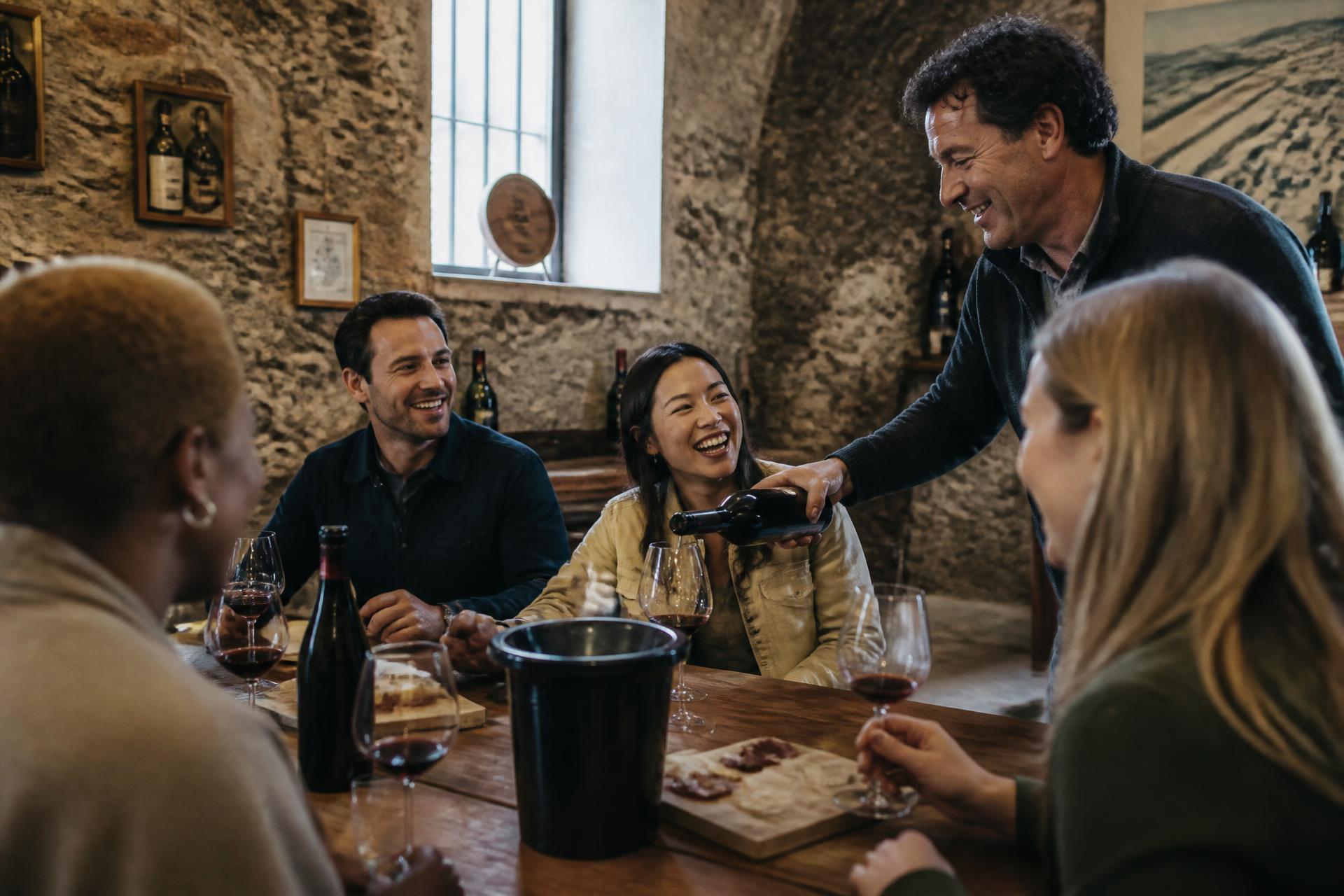 Guests laughing around a table while a host pours wine in a European stone cellar
